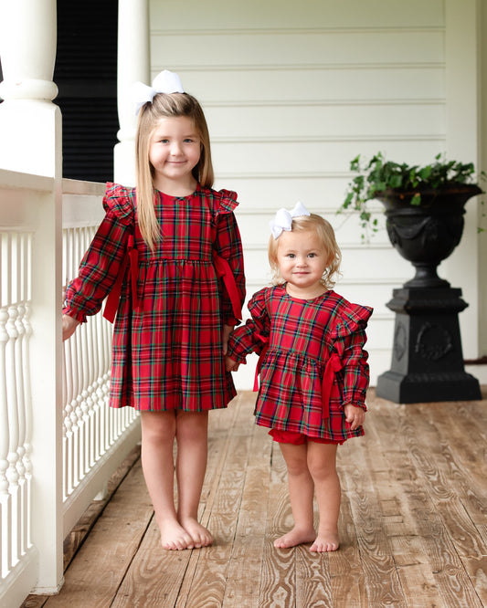 Two young girls in matching red plaid dresses standing on a wooden porch.