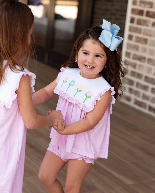 Two young girls in pink dresses with floral details on a wooden deck.