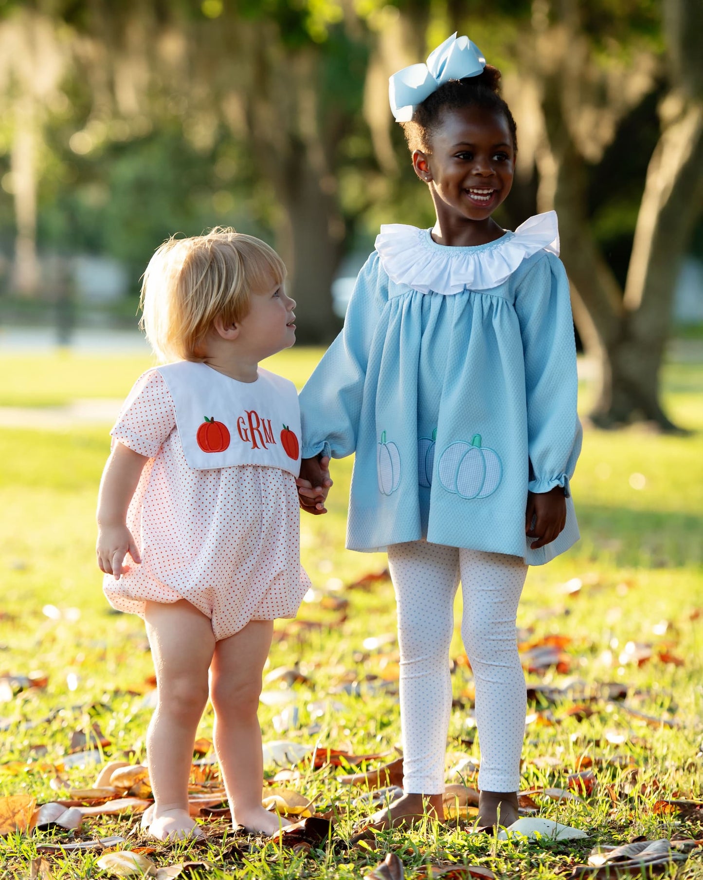 Two young girls in pumpkin outfits standing outdoors with trees in the background