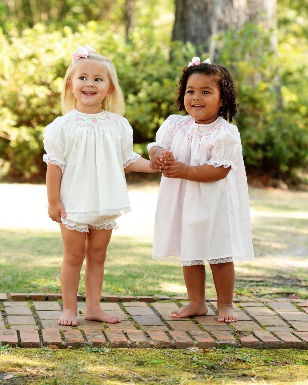 Two young girls in white dresses standing on a brick path with greenery in the background.