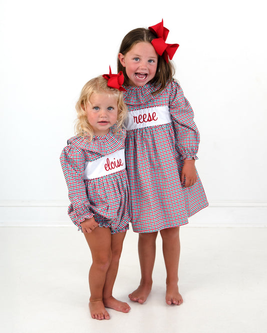 Two young girls modeling matching red, white, and blue gingham dresses with white name patches. The older girl has a large red bow in her hair and the name 'reese' on her chest, while the younger girl has a small red bow and the name 'elise' on her chest.