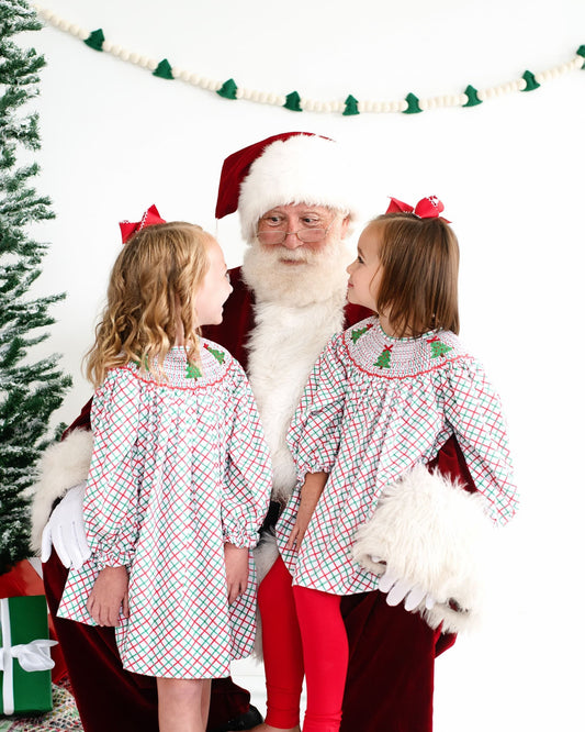 Two young girls sitting with Santa Clause wearing plaid outfits with Christmas trees on them.