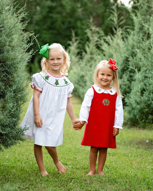 Two young girls standing barefoot on a grassy lawn in front of green trees, wearing matching Christmas-themed dresses. The girl on the left wears a white dress with green embroidered details and a green bow in her hair, and the girl on the right wears a red pinafore-style dress over a white long-sleeve top with a red bow in her hair.