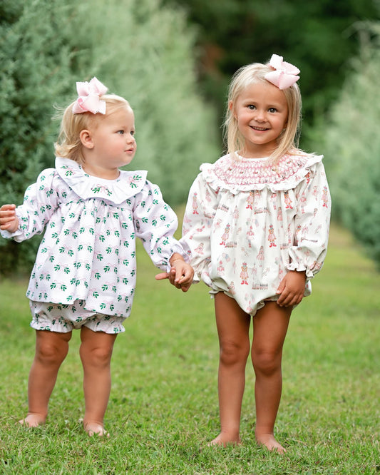 Two young girls standing barefoot on a grassy lawn outdoors, wearing matching white long-sleeve romper outfits with small pink and green floral prints and pink bows in their hair.
