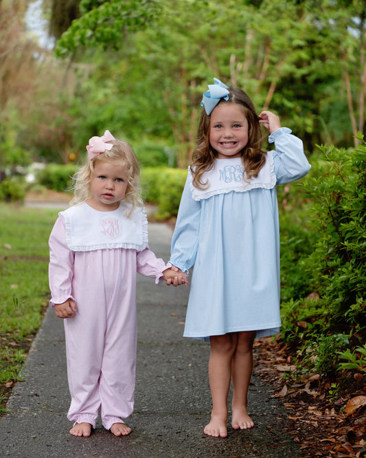 Two young girls standing barefoot on a paved path outdoors, wearing long-sleeve baby dresses. The girl on the left wears a light pink dress with a white ruffled collar and a pink bow in her hair, and the girl on the right wears a light blue dress with a white ruffled collar, blue bow, and monogrammed neckline.