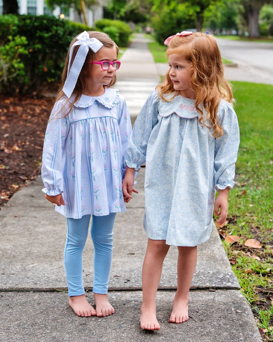 Two young girls standing on a sidewalk outdoors, wearing light blue long-sleeve dresses with white ruffled collars. One dress has a subtle floral print, and the other is solid light blue.