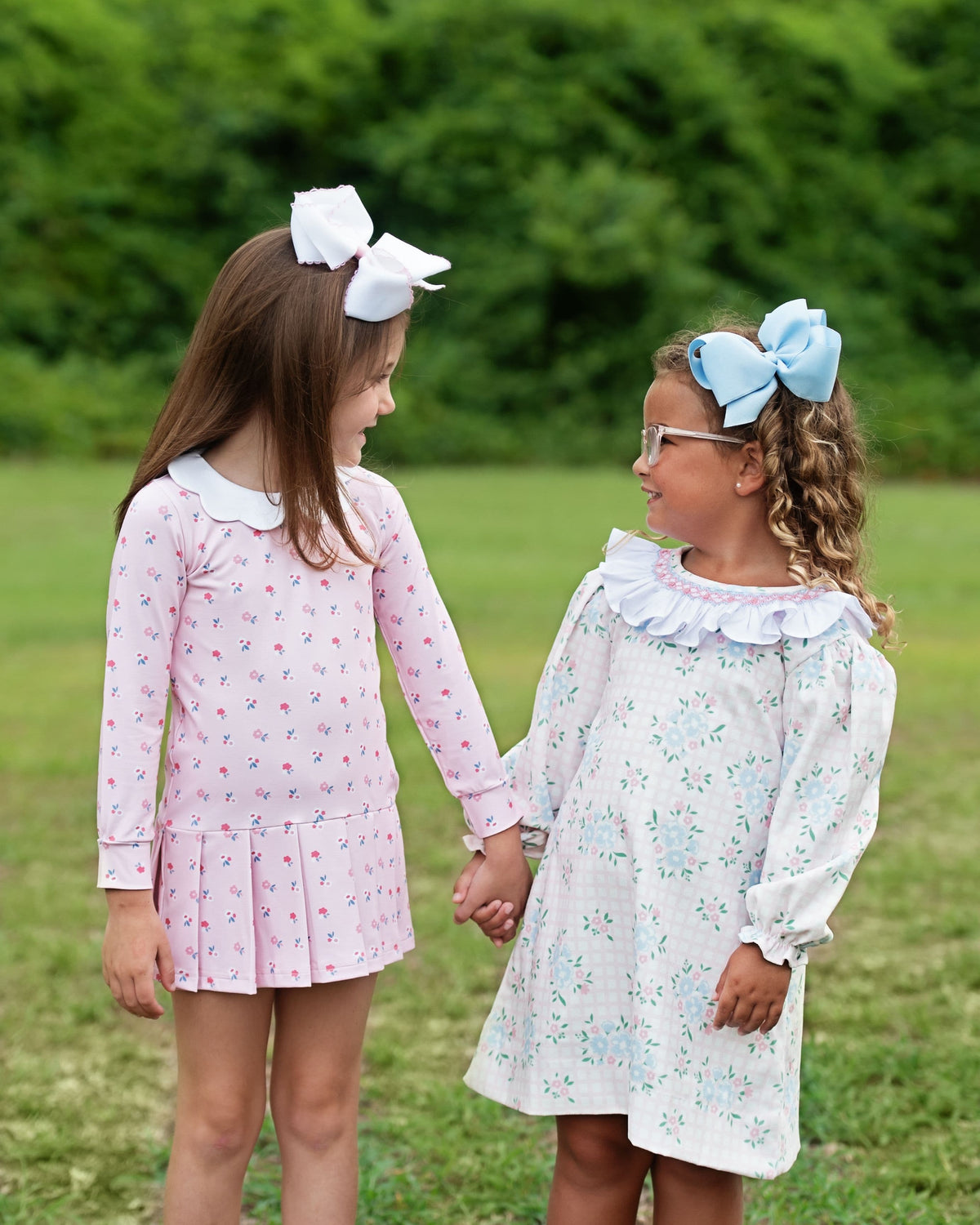 Two young girls standing outdoors in a grassy park holding hands. The girl on the left wears a pink long-sleeve dress with a white Peter Pan collar and a white bow in her hair.