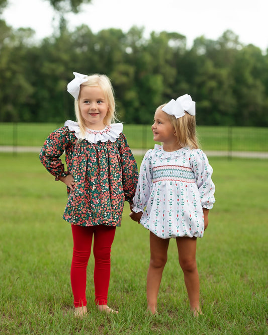 Two young girls standing outdoors on a grassy field, each wearing a long-sleeve floral or patterned dress with a ruffled collar and a large white bow in their hair. The girl on the left wears red leggings, and the girl on the right is barefoot.