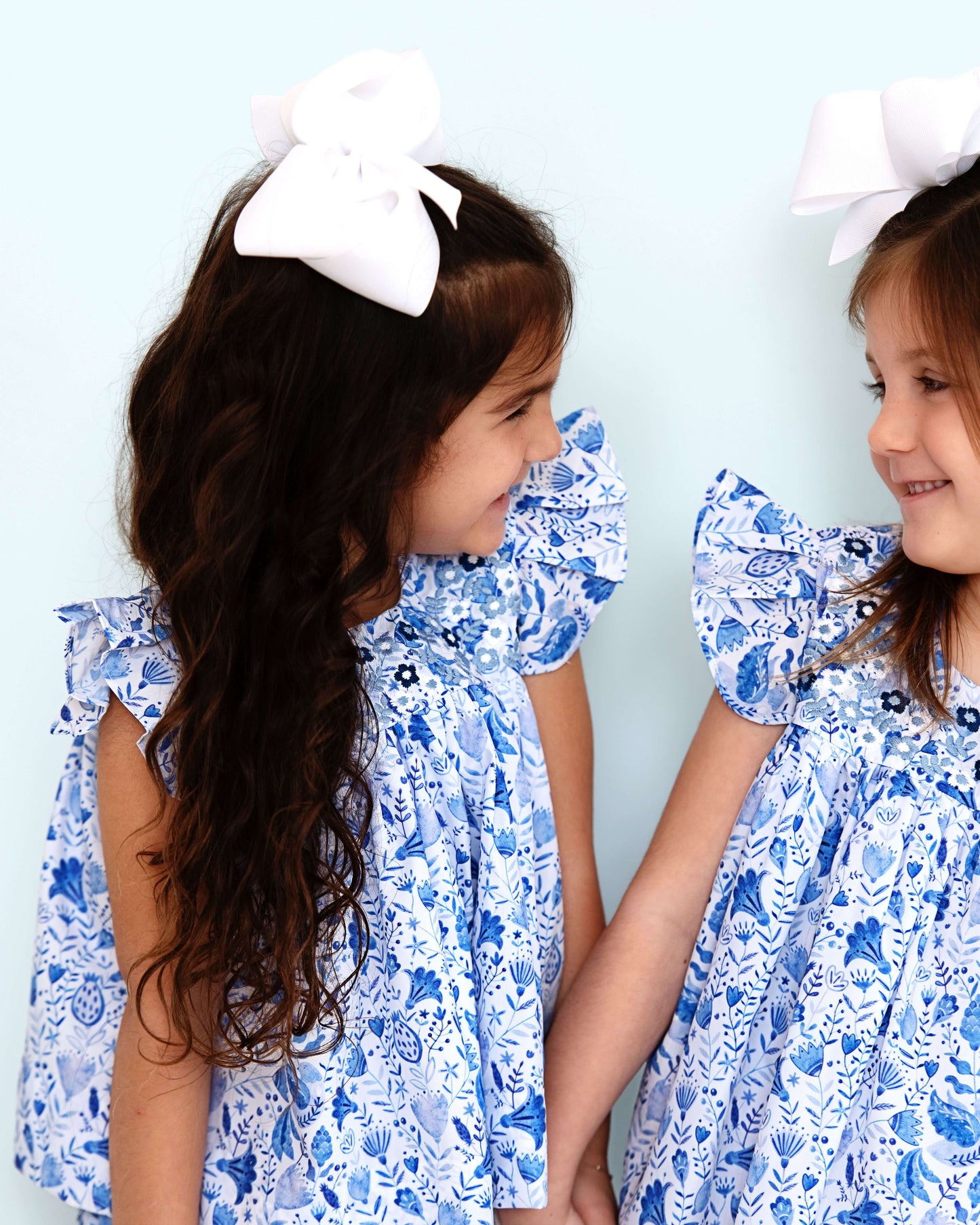 Two young girls wearing blue floral dresses with white bows in their hair against a light blue background.