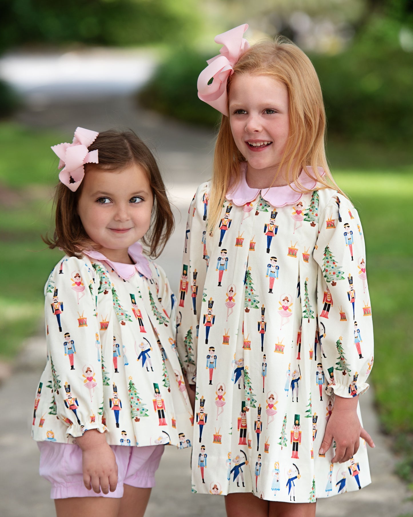 Two young girls wearing matching dresses with a pattern of children playing outdoors, standing on a path with greenery in the background.