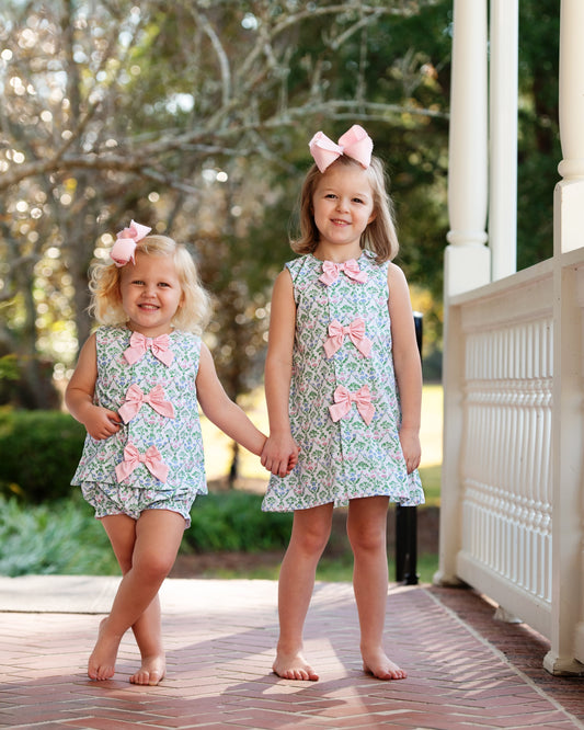 Two young girls wearing matching dresses with pink bows outdoors.
