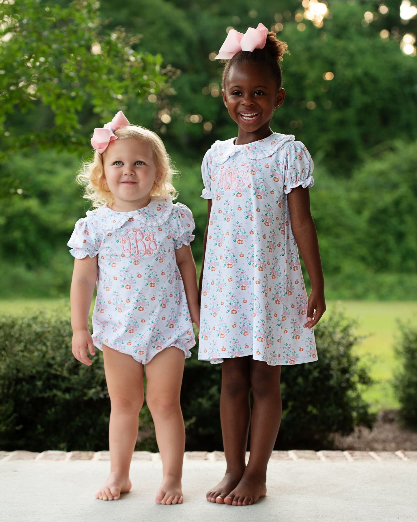Two young girls wearing matching floral dresses outdoors with greenery in the background.