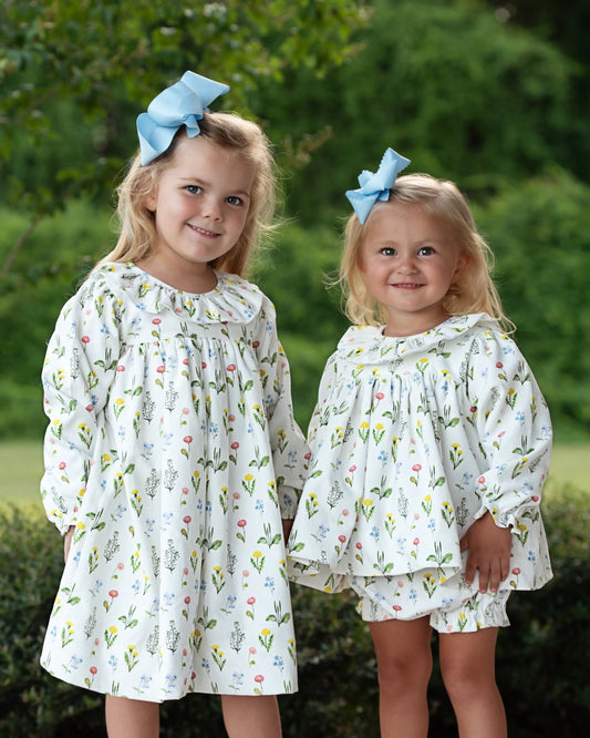Two young girls wearing matching floral outfits outdoors with greenery in the background.