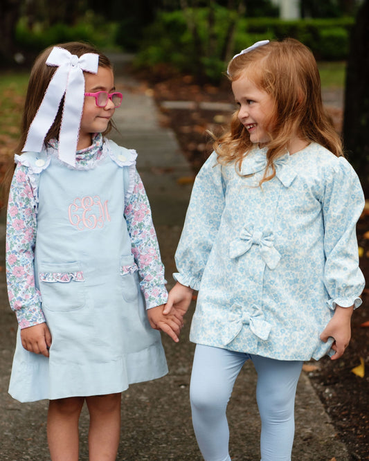 Two young girls wearing matching light blue patterned outfits, standing outdoors on a path and holding hands. One girl wears a light blue dress with a large white bow headband, and the other wears a light blue patterned long-sleeve top with matching light blue leggings.