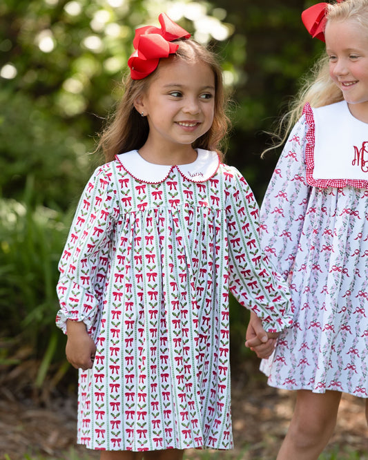 Two young girls wearing matching long-sleeve dresses with a white base and red bow pattern, standing outdoors in a garden setting. The girl on the left wears a large red bow in her hair, and both girls are smiling and holding hands.