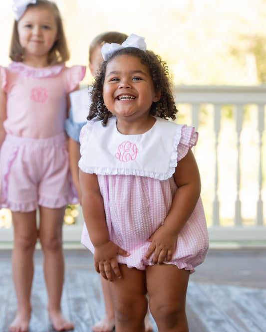 Two young girls wearing matching pink seersucker outfits with white ruffled collars on a wooden deck.