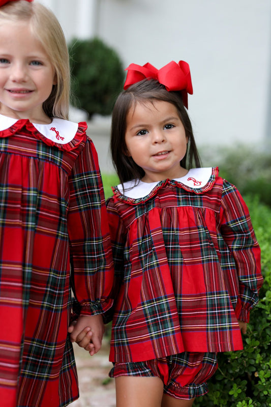 Two young girls wearing matching red plaid dresses with white Peter Pan collars, standing outdoors and holding hands. The girl on the right also wears a large red bow in her hair.