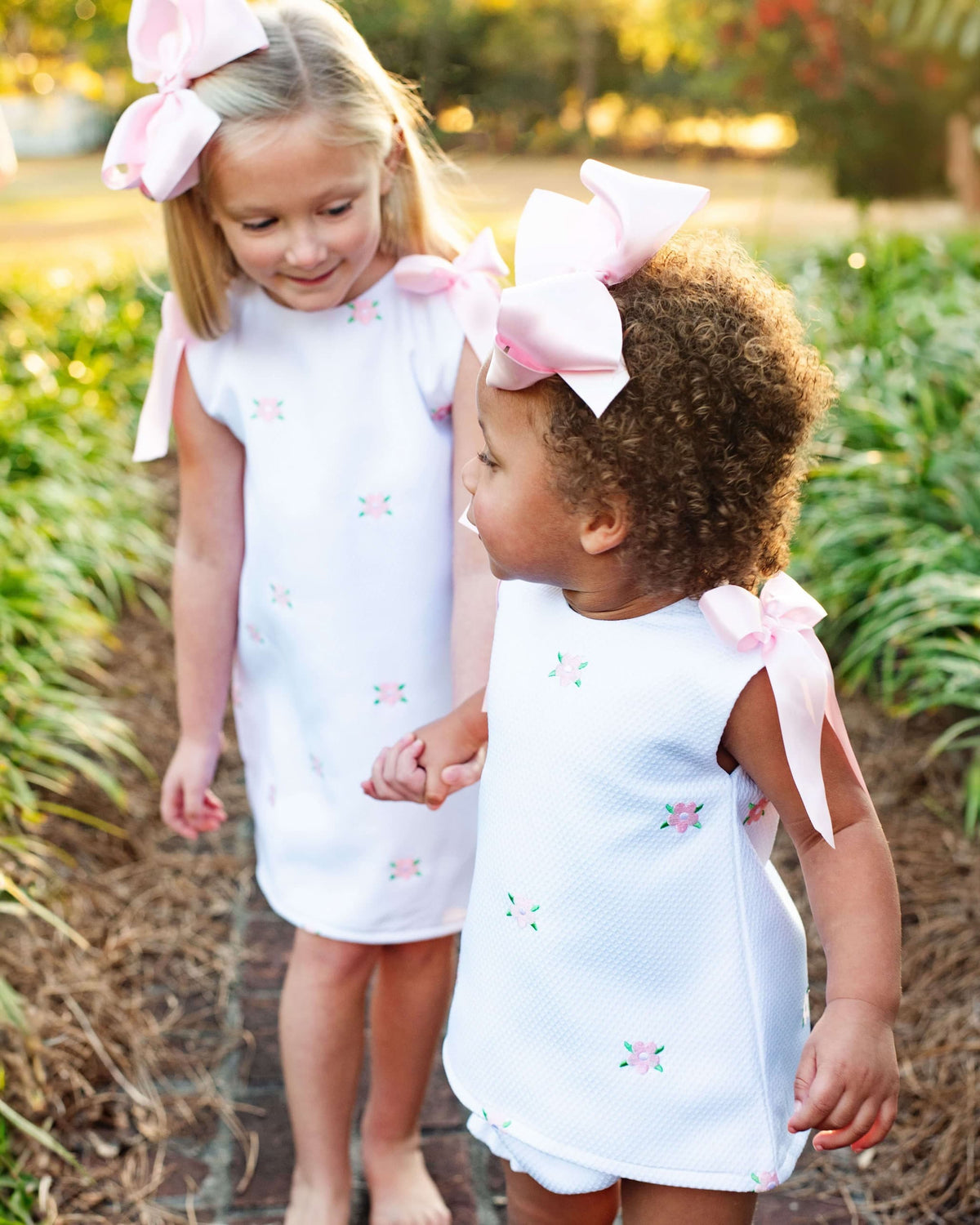 Two young girls wearing matching white outfits with embroidered flowers and pink bows