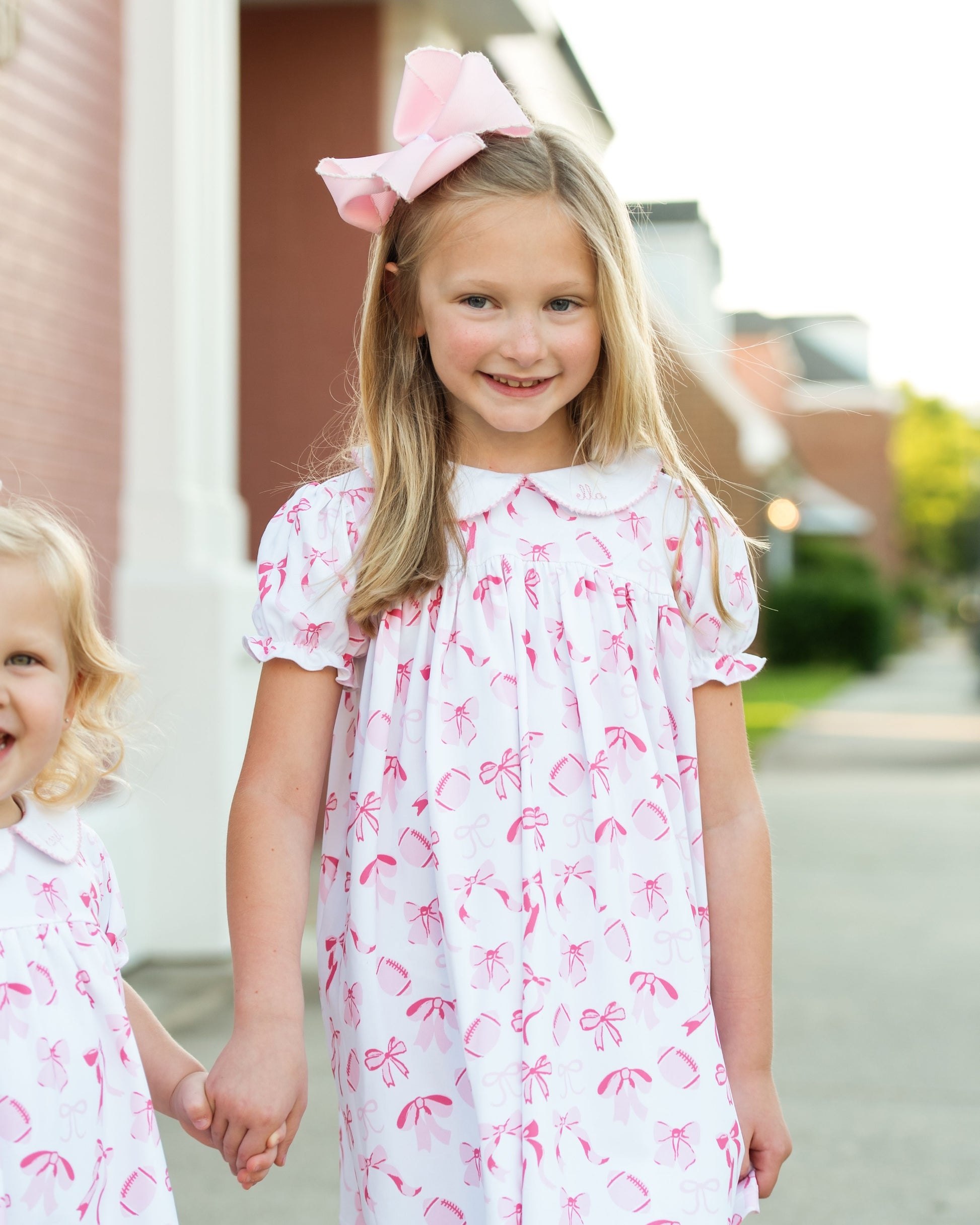 Two young girls wearing white dresses with pink bow patterns, standing outdoors.