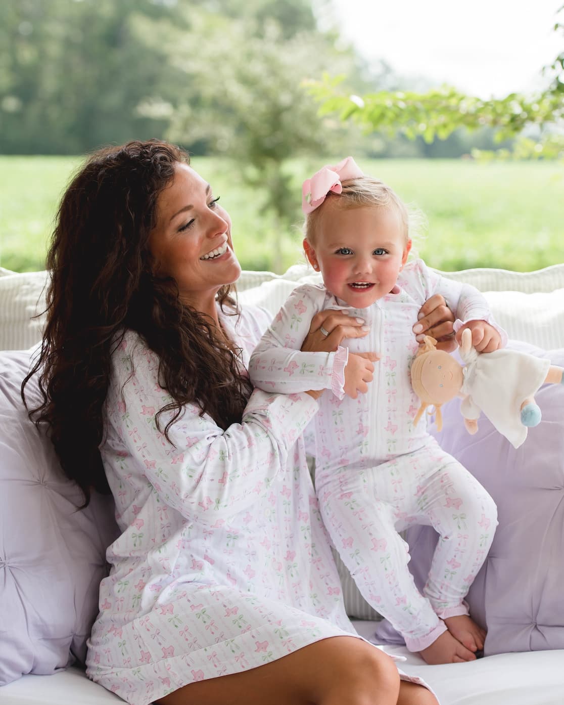 Woman holding a baby in a white outfit with a pink bow outdoors.