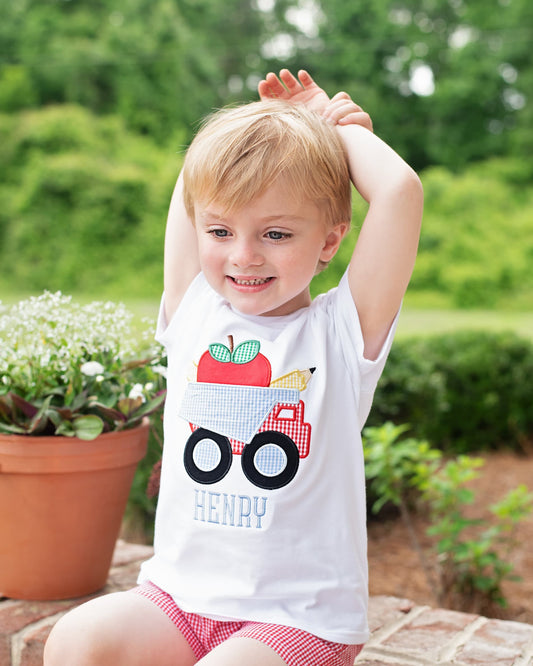 Young boy holding hands over his head. smiling, wearing a school themed shirt with a dump truck, apple and pencil.