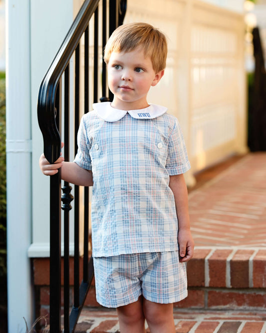 Young boy in a blue checkered outfit standing on a porch.