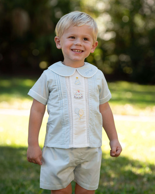 Young boy in a light-colored outfit standing outdoors with greenery in the background
