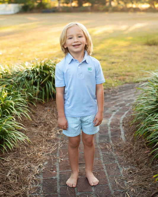 Young boy in a light blue polo shirt and shorts standing on a brick path with greenery around.