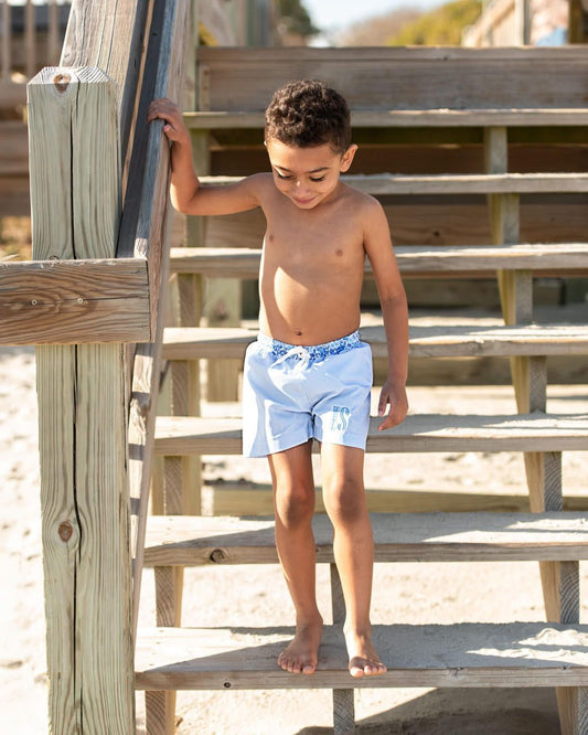 Young boy in light blue swim shorts standing on a wooden staircase.