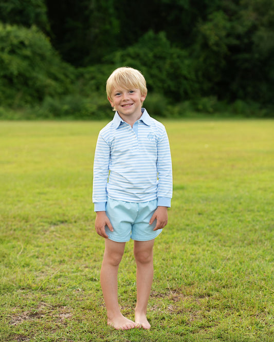 Young boy standing in a grassy field wearing a striped shirt and light shorts.