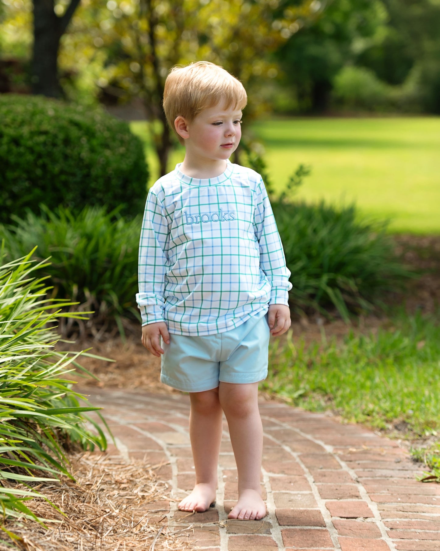 Young boy standing on a brick path in a garden wearing a checkered shirt and light shorts.
