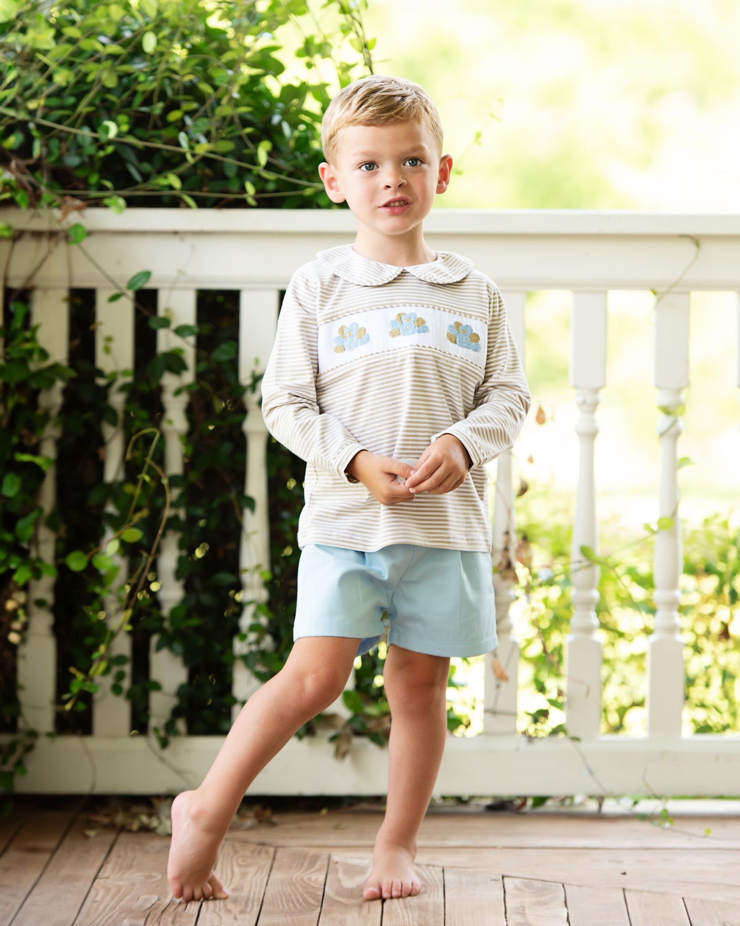 Young boy standing on a wooden deck wearing a striped shirt and light blue shorts.