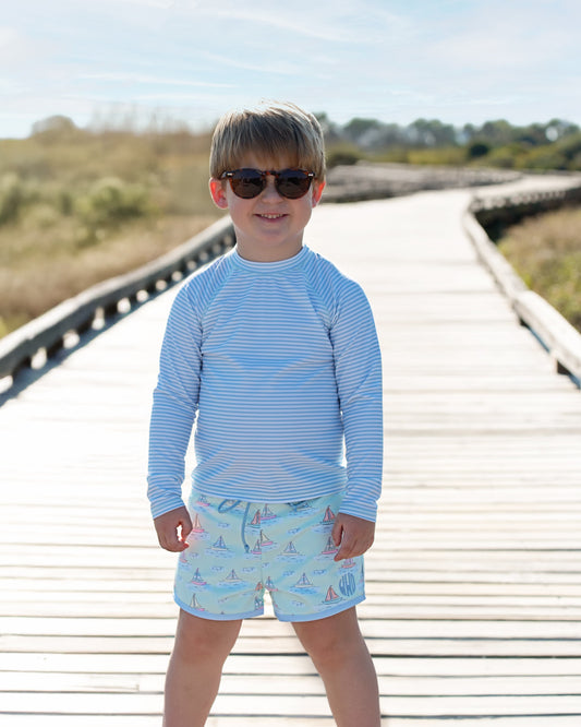 Young boy wearing a blue striped rash guard and swim shorts on a wooden boardwalk.
