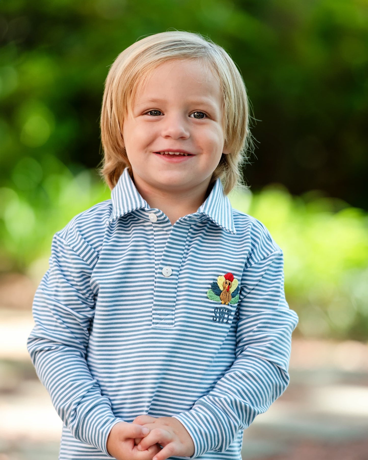Young boy wearing a blue striped right with a logo standing outdoors with greenery in the background