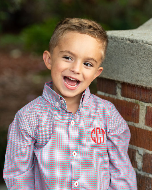 Young boy wearing a checkered shirt with a monogram, standing in front of a brick wall.
