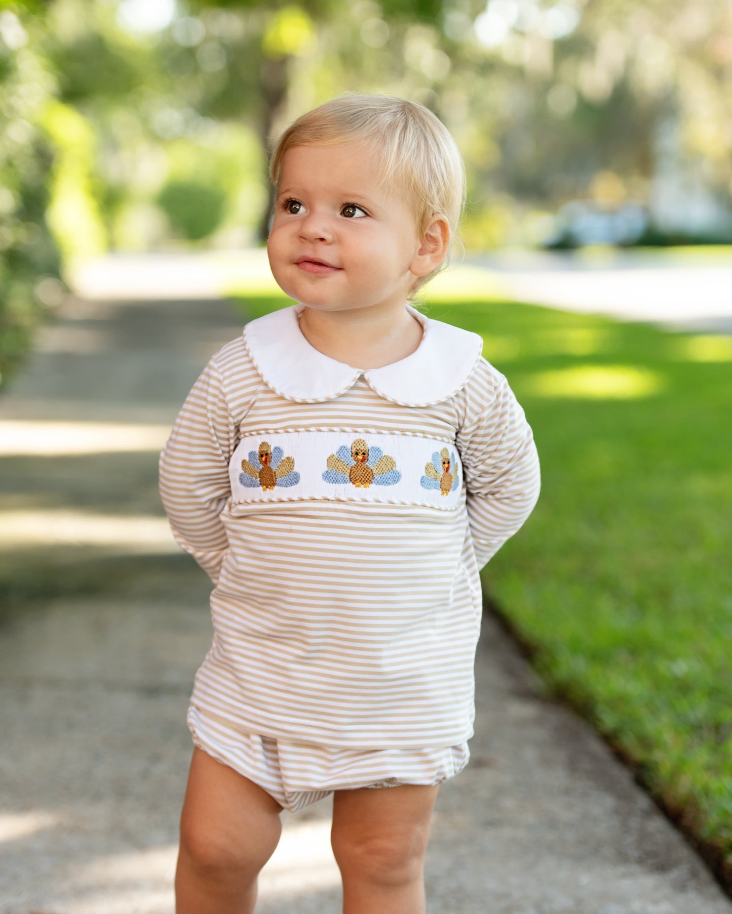 Young boy wearing a khaki striped diaper set with a collar and turkeys outdoors
