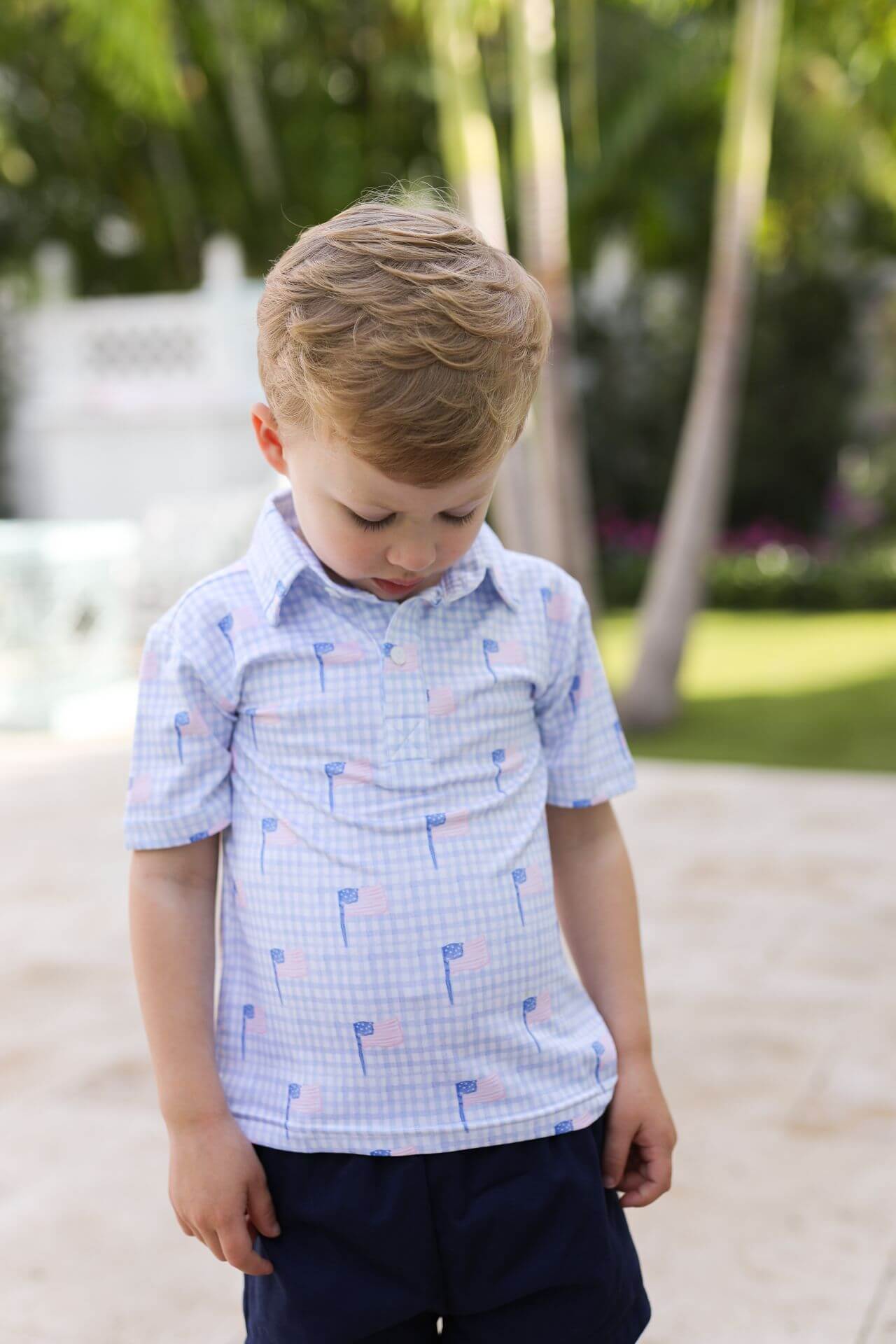 Young boy wearing a light blue checkered shirt with small patterns, standing outdoors with greenery in the background.
