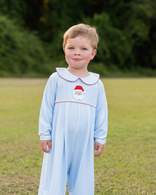 Young boy wearing a light blue dress with a red collar and Santa hat design, standing outdoors.