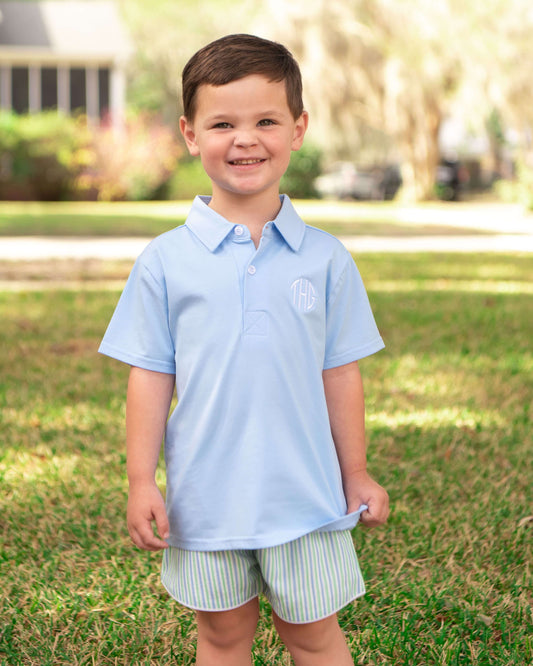 Young boy wearing a light blue polo shirt and striped shorts standing on grass.
