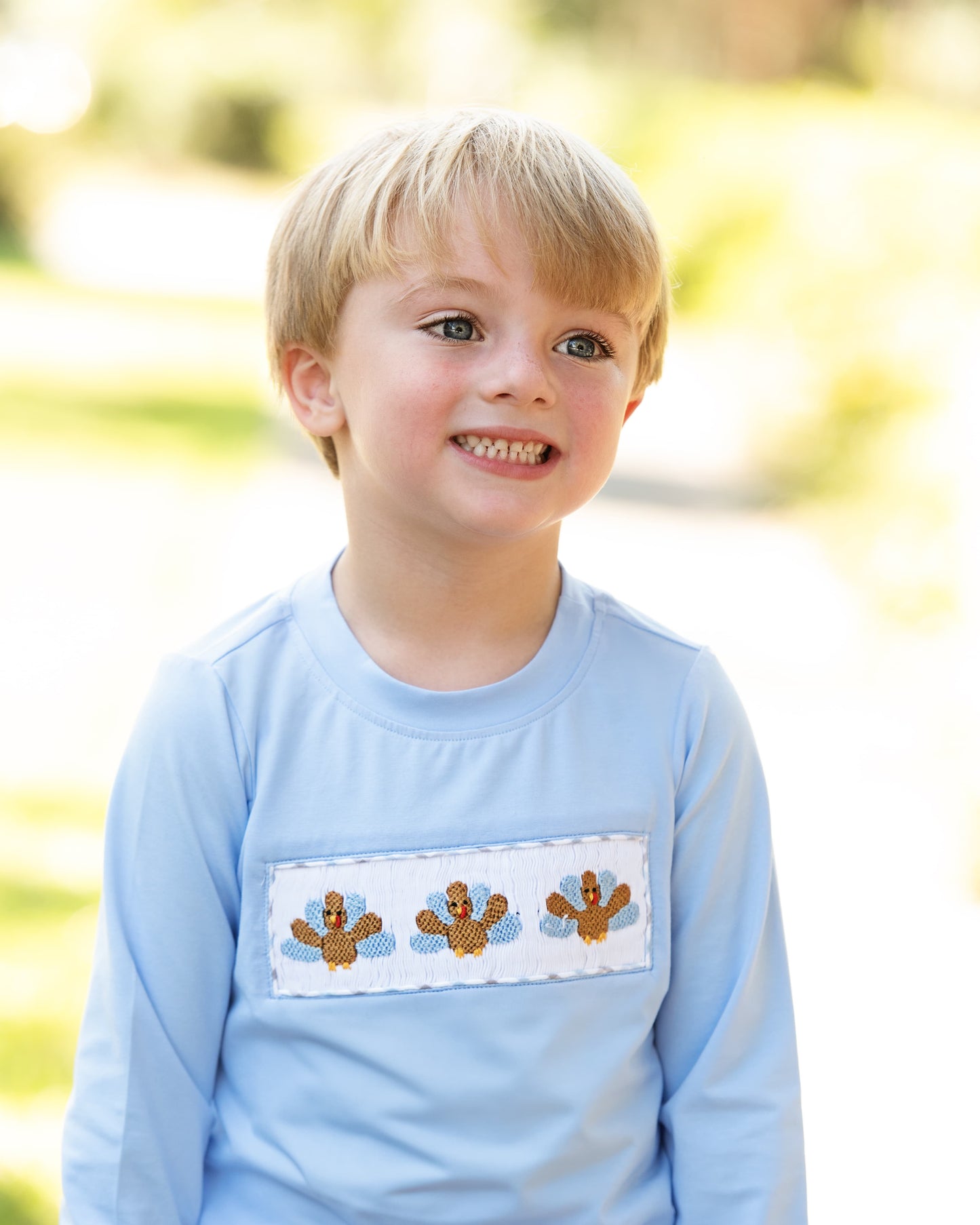 Young boy wearing a light blue shirt with smocked turkeys design outdoors.