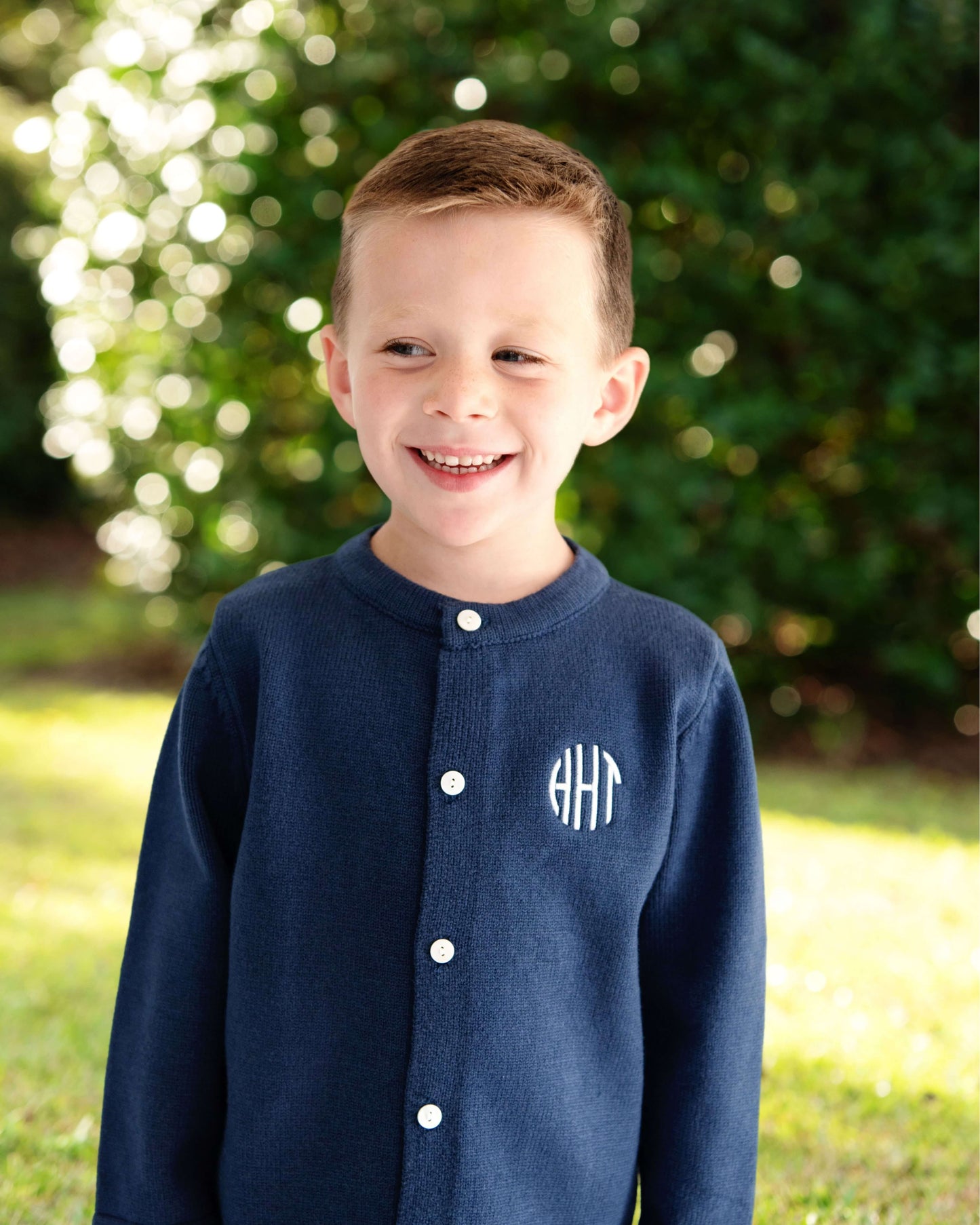 Young boy wearing a navy blue jacket with a monogram, standing outdoors with greenery in the background.
