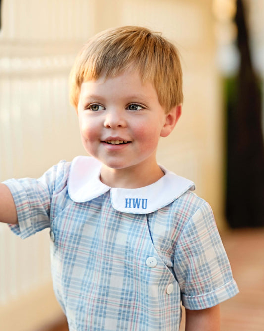 Young boy wearing a plaid shirt with a white collar and 'HWU' embroidered on it, standing outdoors.