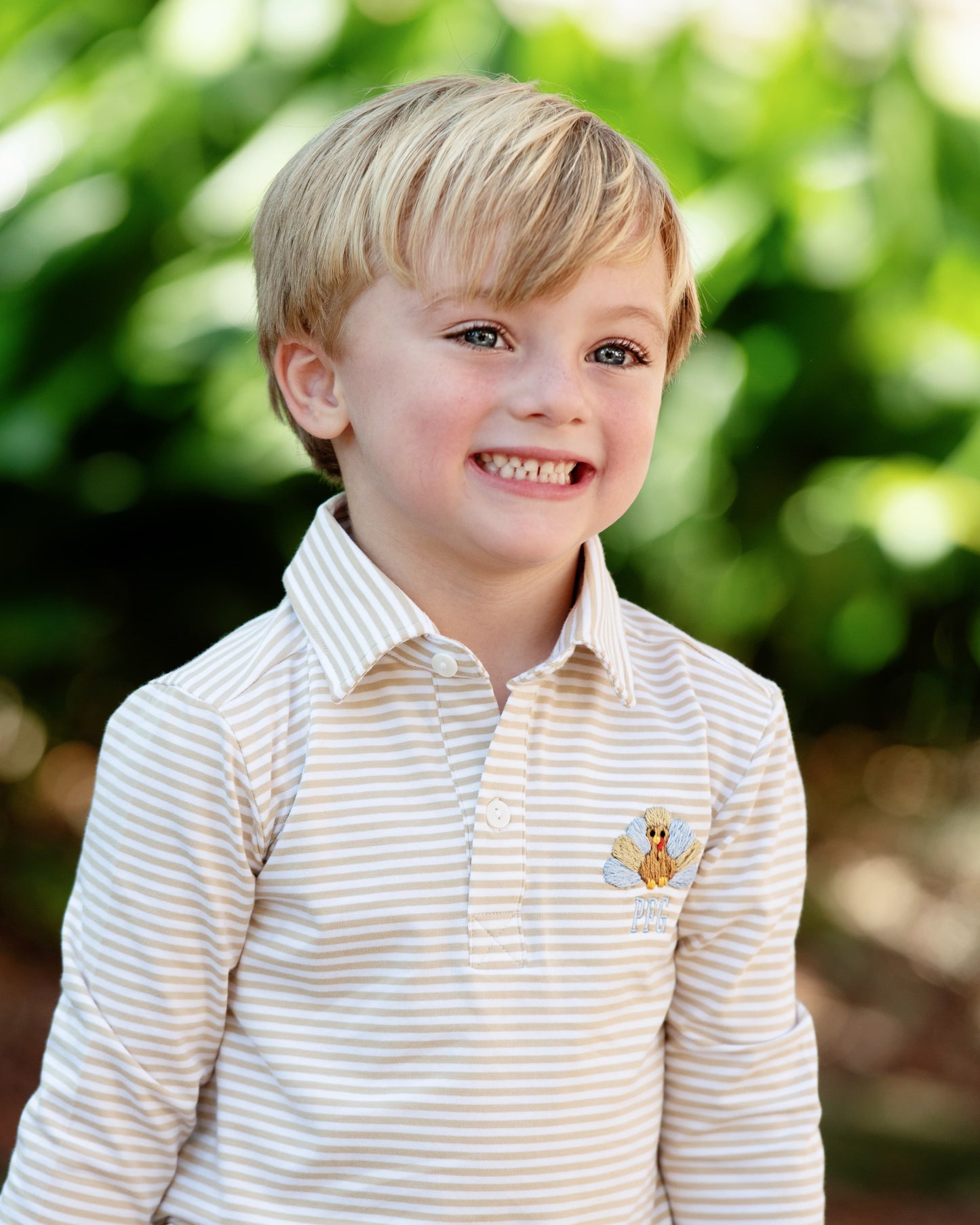 Young boy wearing a striped knit polo shirt with a small embroidered turkey, standing outdoors with greenery in the background.