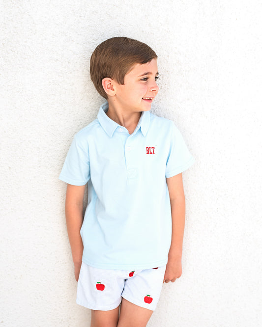 Young boy wearing blue poo shirt and blue gingham shorts with apples embroidered.