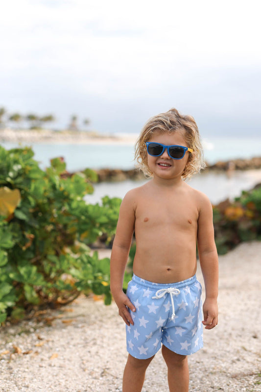 Young boy wearing blue swim shorts with a star pattern, standing on a beach.