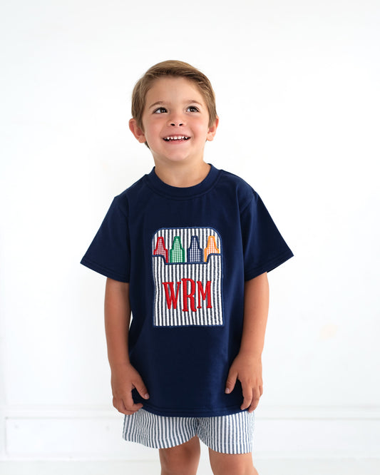 Young boy wearing navy outfit with crayon box on shirt.