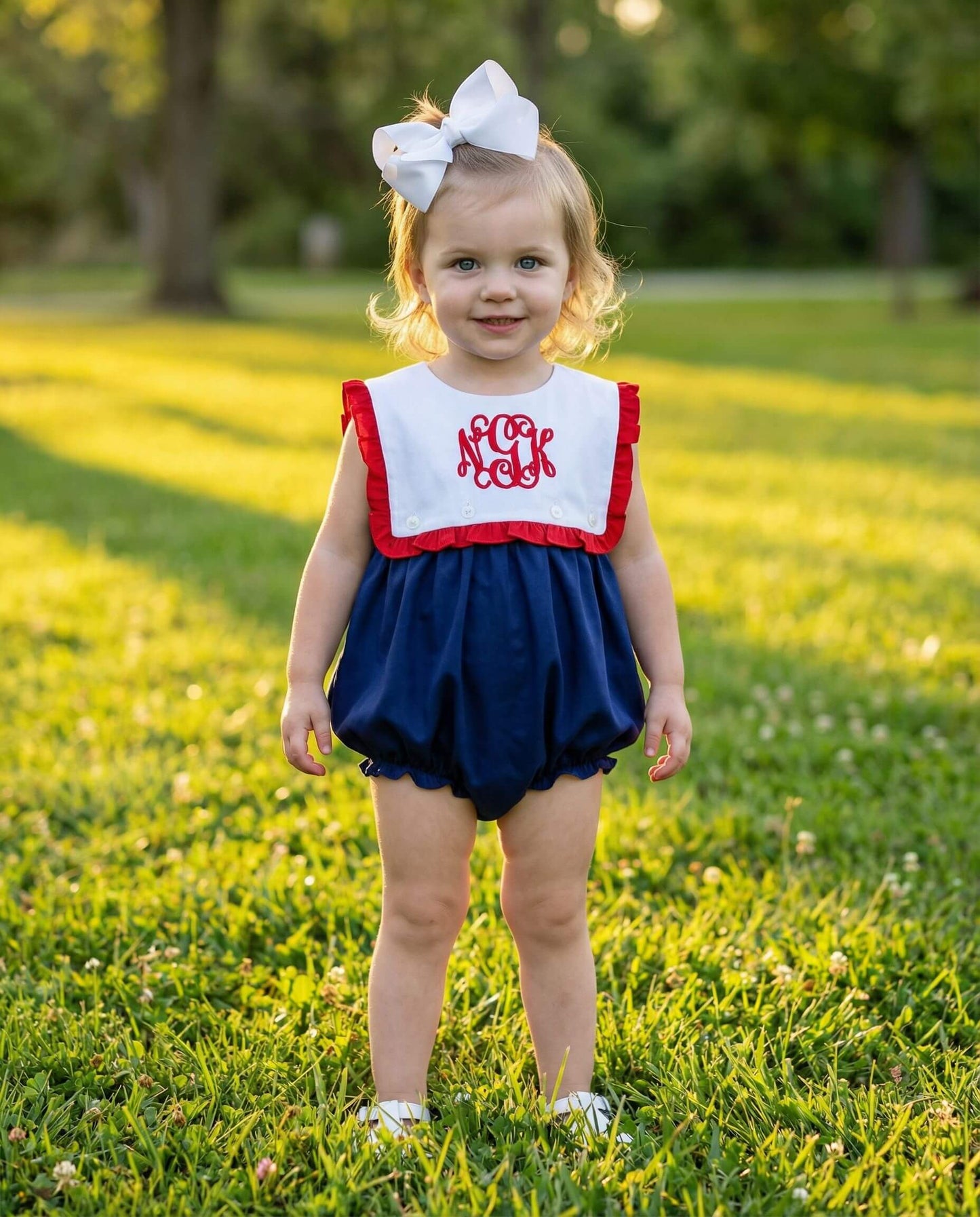Young child wearing a red, white, and blue outfit with a monogrammed top in a grassy outdoor setting.