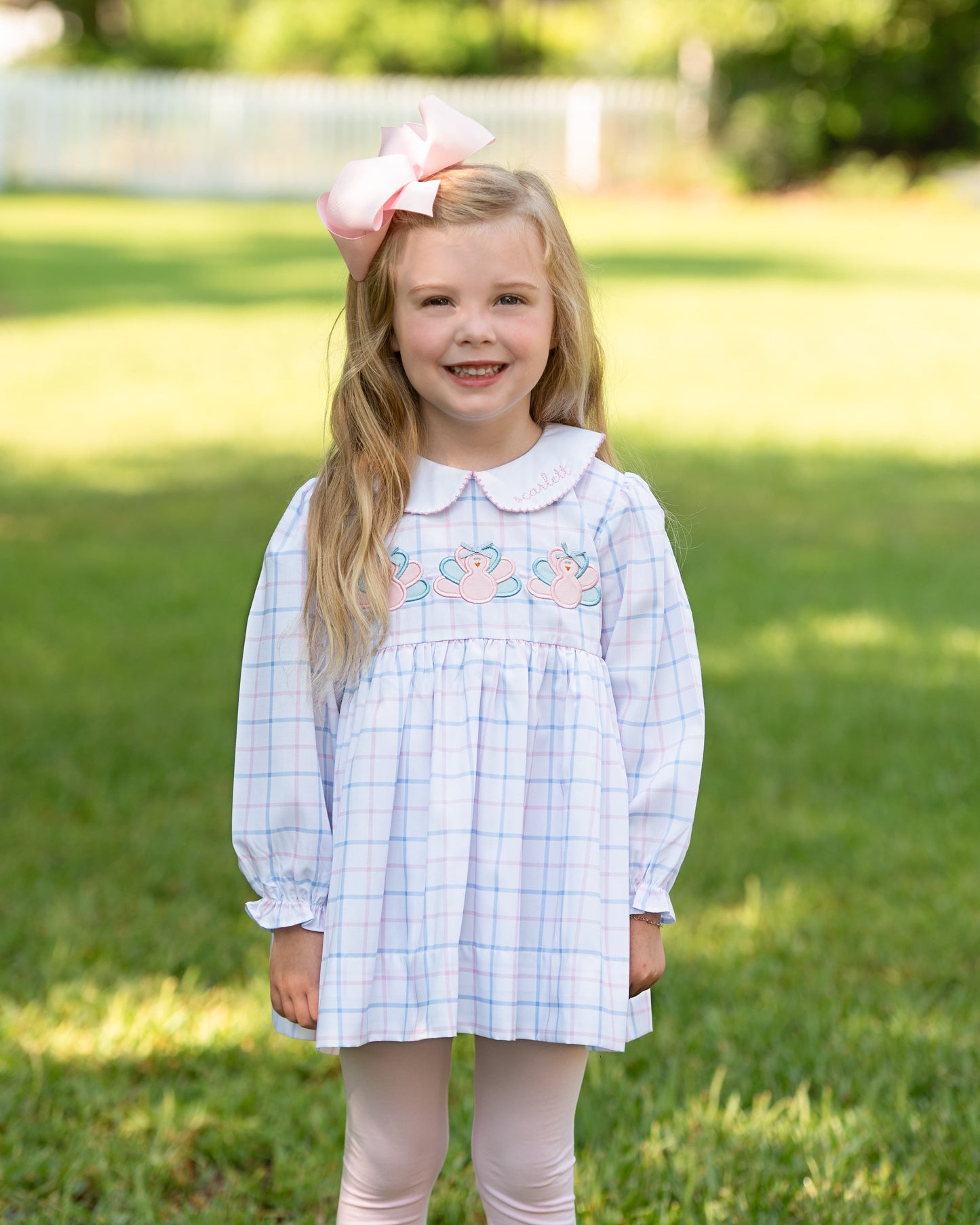 Young girl in a blue checkered dress with floral embroidery standing on grass.