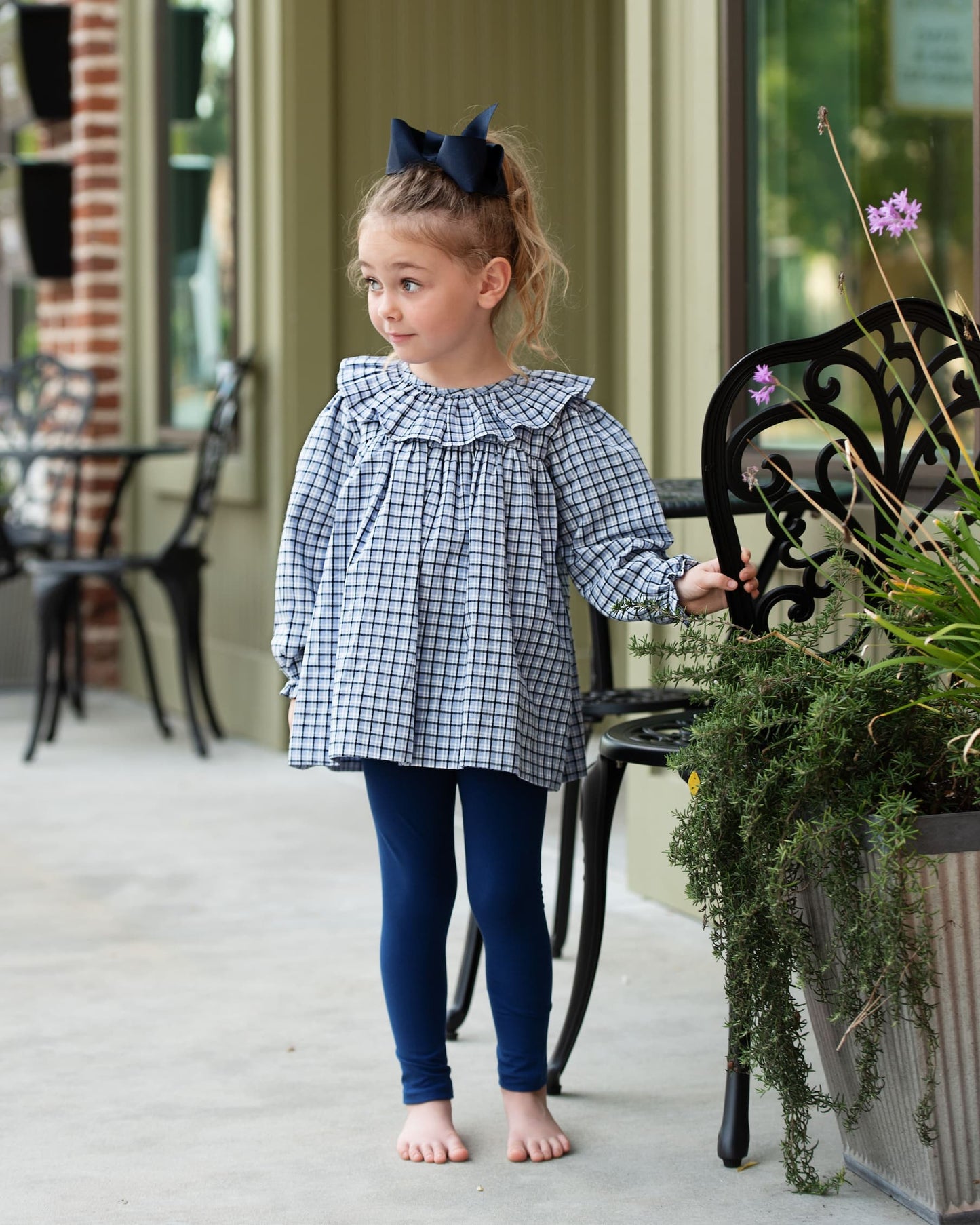 Young girl in a checkered dress and blue leggings standing on a porch.