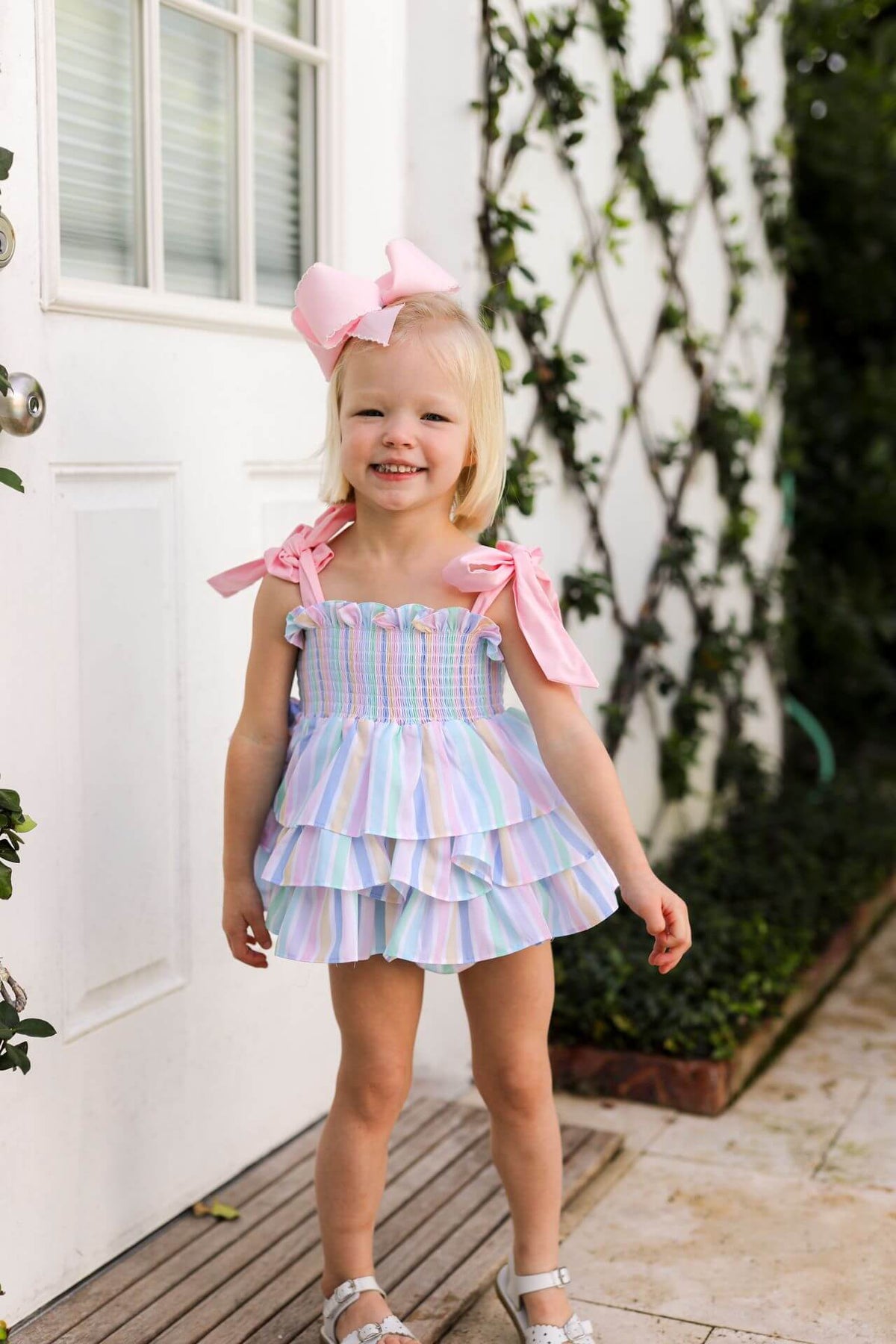 Young girl in a colorful dress with pink bows standing in front of a white door.